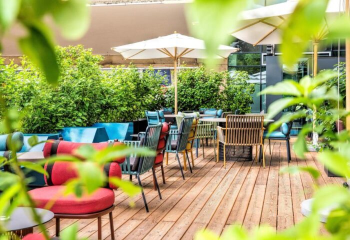Motel One Wien Westbahnhof's outdoor patio. Gay-friendly hotel in Vienna with tables, chairs, umbrellas, and greenery on a wooden deck.
