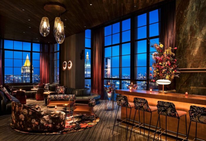 Moxy NYC Chelsea gay-friendly hotel bar. City views from floor-to-ceiling windows. Stylish seating, bar stools, and floral arrangements.