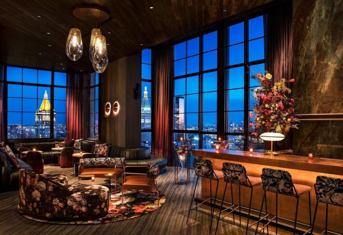 Moxy NYC Chelsea gay-friendly hotel bar. City views from floor-to-ceiling windows. Stylish seating, bar stools, and floral arrangements.
