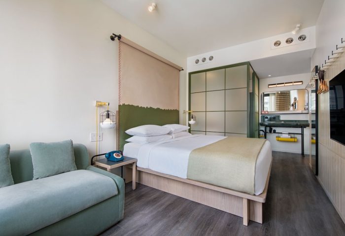 Moxy NYC Chelsea gay-friendly hotel room. Queen bed, blue couch, and retro phone. Modern design with a view of the bathroom area.
