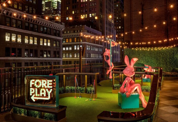 Rooftop mini golf at the gay-friendly Moxy NYC Times Square Hotel. Quirky statues and city views. Fore Play sign glows. Night scene.