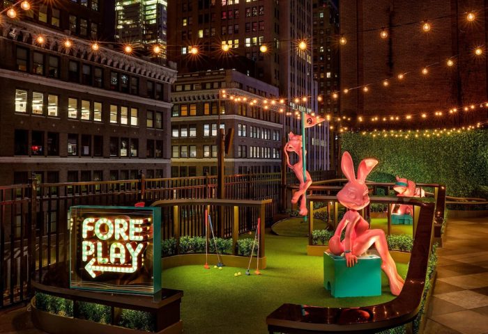 Rooftop mini golf at the gay-friendly Moxy NYC Times Square Hotel. Quirky statues and city views. Fore Play sign glows. Night scene.