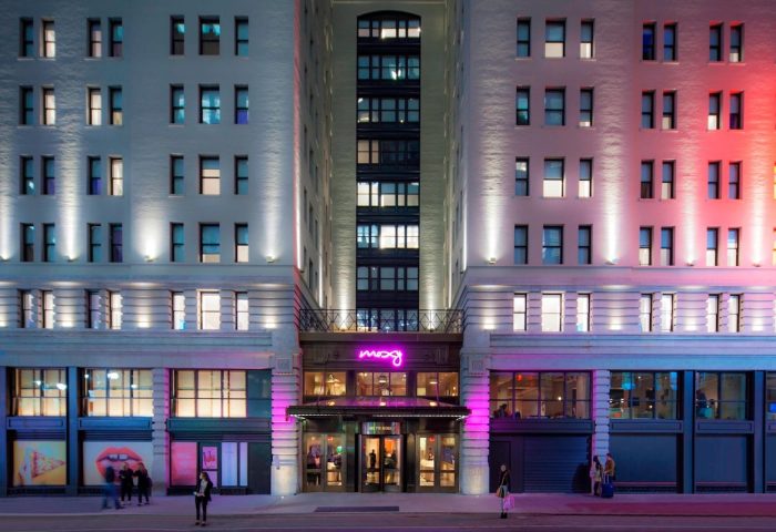 Exterior of Moxy NYC Times Square, a gay-friendly hotel. The building is lit with pink and red lights. Neon sign above the entrance.