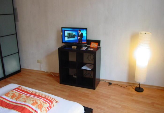 My Gayhostel room interior: TV atop a black shelf, next to a lamp. Gay-friendly hotel room featuring a bed and modern amenities for gay travelers.