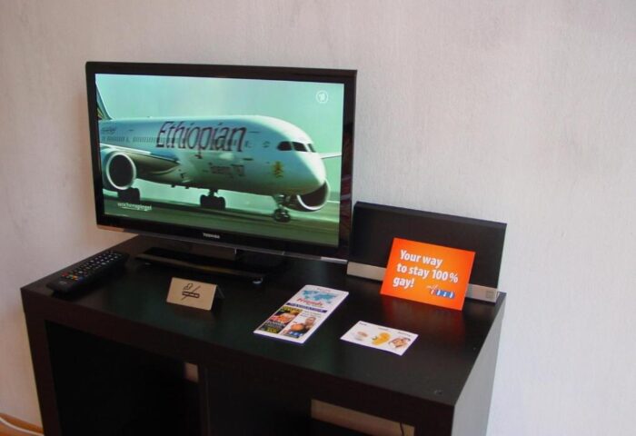 Gay-friendly hotel room at My Gayhostel: TV showing an Ethiopian Airlines plane, remote, and a sign that says Your way to stay 100% gay!.