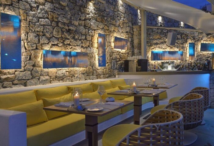 Myconian Korali Relais & Chateaux gay-friendly hotel restaurant. Stone wall, yellow seating, tables set with candles. Upscale dining experience.