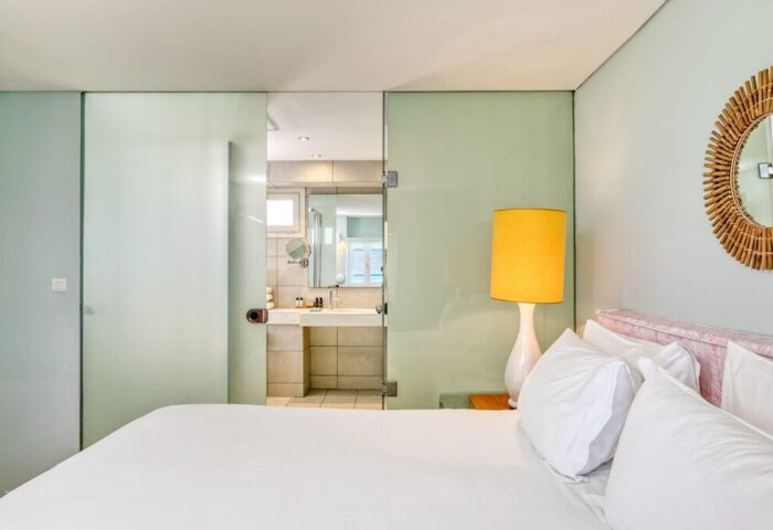 Mykonos Theoxenia gay-friendly hotel room. View of bed, glass wall to bathroom, yellow lamp, & round mirror. Modern design for gay travelers.