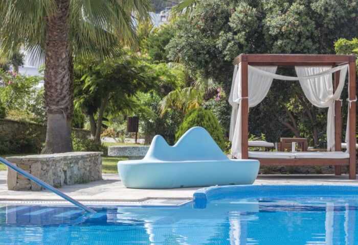 Mykonos Theoxenia gay-friendly hotel pool view. Stylish blue seating, cabana, palm trees, and crystal clear water. Ideal gay travel destination.