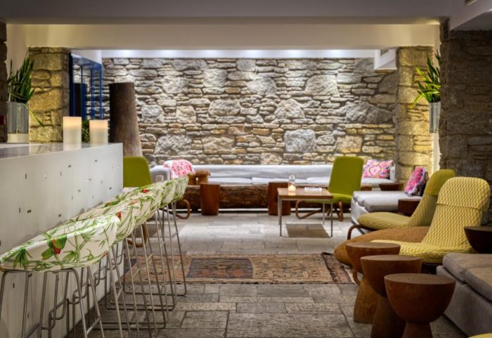 Mykonos Theoxenia, a gay-friendly hotel. Interior view with stone wall, stylish seating, and bar stools. Ideal for gay travel in Mykonos.