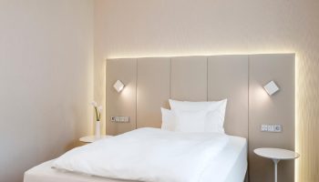 NH Collection Frankfurt City hotel room. Modern design with white linens, side tables, and sleek lighting. Gay-friendly hotel for travelers.