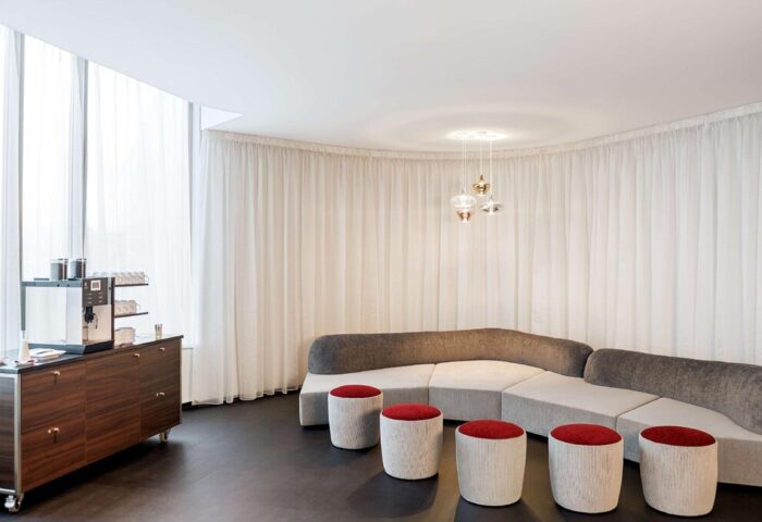 NH Collection Frankfurt City gay-friendly hotel lounge area. Modern design with curved sofa, red stools, coffee station, and sheer curtains.