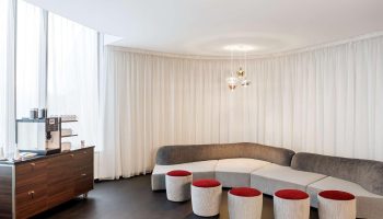 NH Collection Frankfurt City gay-friendly hotel lounge area. Modern design with curved sofa, red stools, coffee station, and sheer curtains.