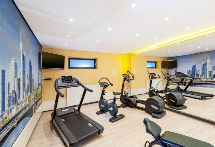 Gym at NH Collection Frankfurt City, a gay-friendly hotel. Treadmill, exercise bike, and elliptical machine. Frankfurt skyline mural on the wall.