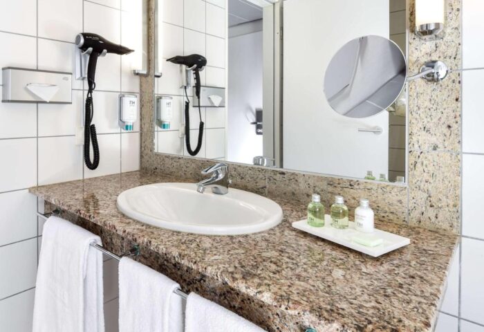 NH Collection Frankfurt City gay-friendly hotel bathroom. Sink, granite countertop, mirror, hair dryer, amenities, and white towels.