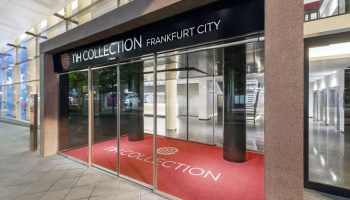 Entrance to the gay-friendly NH Collection Frankfurt City Hotel. Red carpet with the hotel logo welcomes guests. Modern architecture.