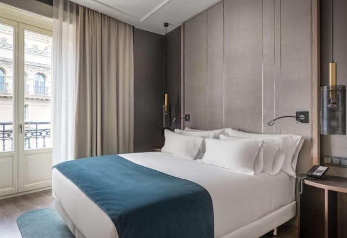 NH Collection Madrid Gran Via gay-friendly hotel room with a large bed, crisp white linens, and a blue throw. Pet-friendly. Balcony view.