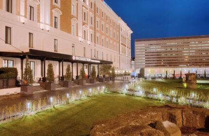 NH Collection Palazzo Cinquecento hotel exterior at night. Gay-friendly hotel with modern architecture, garden, and illuminated walkway. Rome travel.