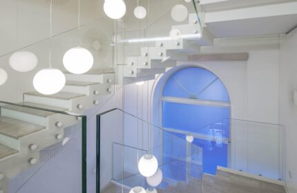 Staircase at NH Milano Palazzo Moscova, a gay-friendly hotel. Modern design with white steps, glass railings, and globe lights. Blue window visible.