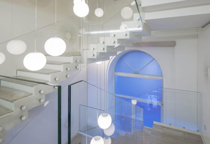 Staircase at NH Milano Palazzo Moscova, a gay-friendly hotel. Modern design with white steps, glass railings, and globe lights. Blue window visible.