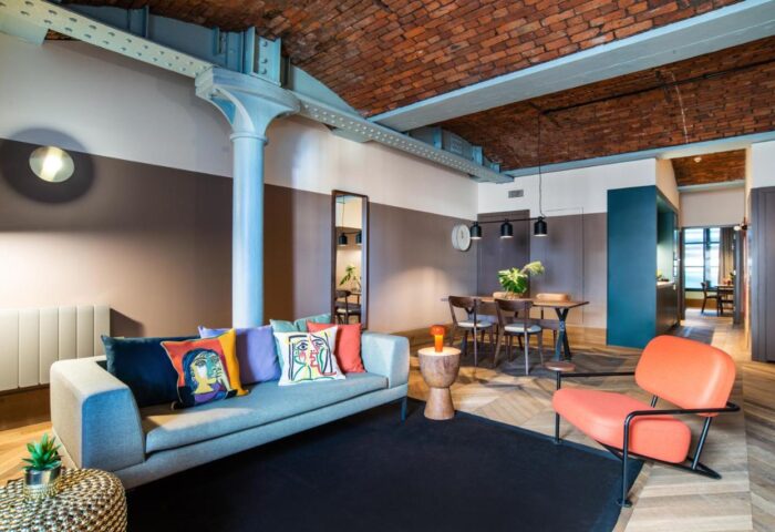 Native Manchester gay-friendly hotel suite. Living area with sofa, colorful pillows, brick ceiling, dining table, and orange chair.