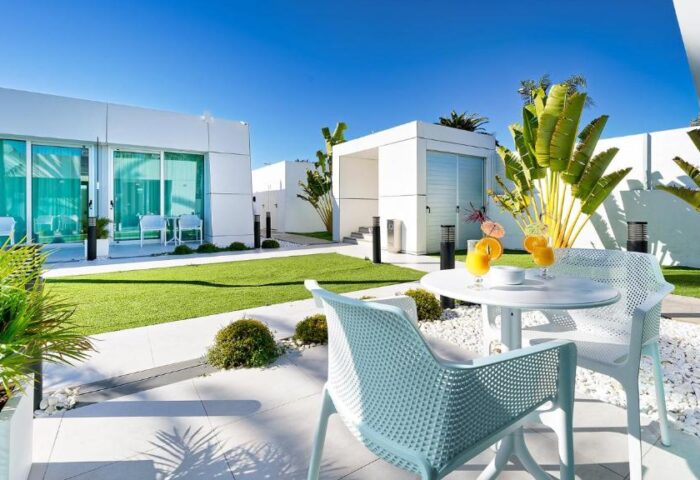 Hotel Nayra Adults Only gay-friendly resort. Outdoor patio with white table, chairs, and drinks. Modern architecture, green lawn, blue sky.