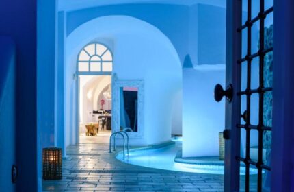 Nefeles Luxury Suites gay-friendly hotel pool view. Blue-lit indoor pool, arched doorways, and stone floors. Luxury gay travel destination.