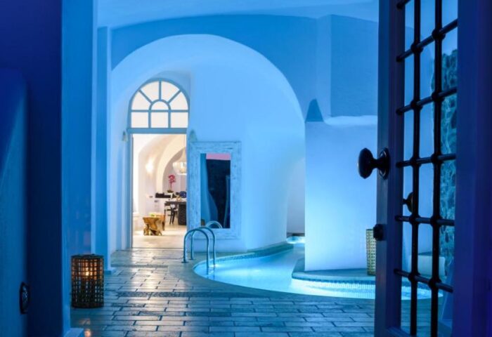 Nefeles Luxury Suites gay-friendly hotel pool view. Blue-lit indoor pool, arched doorways, and stone floors. Luxury gay travel destination.