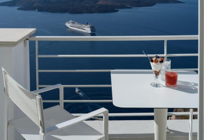 Nefeles Luxury Suites gay-friendly hotel balcony view. Cruise ship on the sea. Dessert and drink on table. Gay travel advice.