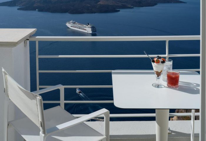 Nefeles Luxury Suites gay-friendly hotel balcony view. Cruise ship on the sea. Dessert and drink on table. Gay travel advice.