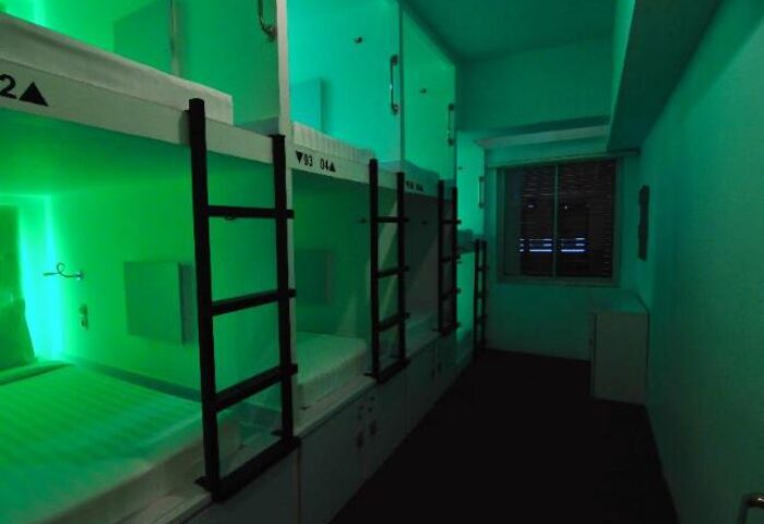 Interior of New Batavia Capsule Hotel, a gay-friendly hotel. Bunk beds with ladders line the wall, lit with green light. Window visible.