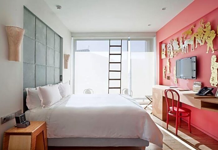 Gay-friendly New Hotel room with modern decor, white bedding, pink accent wall with golden figures, and a ladder leading to a rooftop.