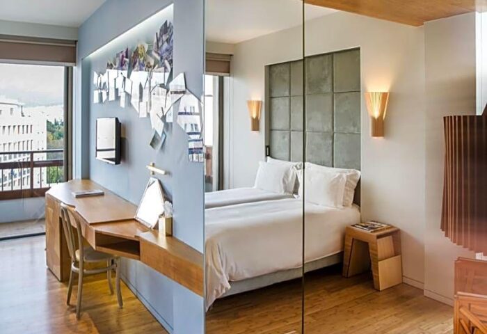 Gay-friendly New Hotel room featuring a desk, TV, and decor on one side, and a bed with a padded headboard and bedside table on the other.