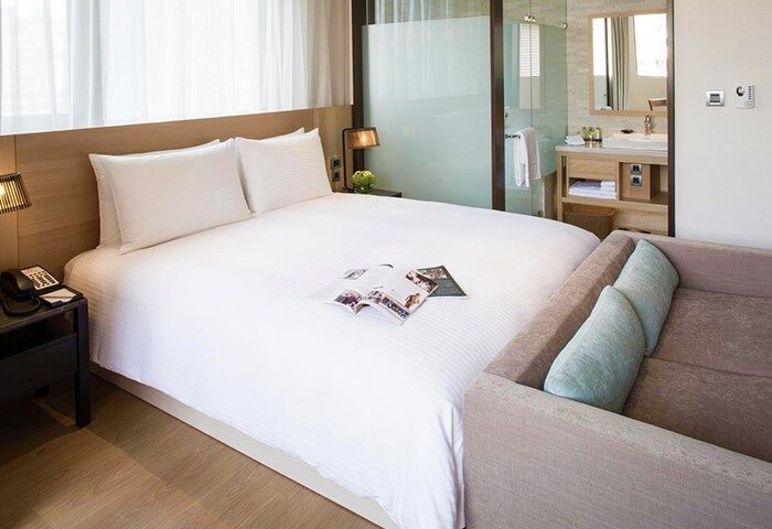 New Riverview Suites gay-friendly hotel room. Bed with white linens, open magazine, and a small couch. Bathroom visible through glass wall.