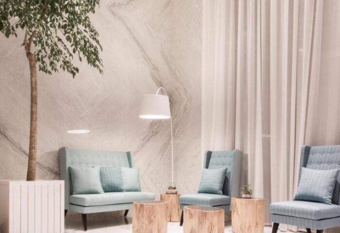 Lobby seating area at New Riverview Suites, a gay-friendly hotel. Light blue chairs, wood tables, tree, and lamp create a welcoming space.