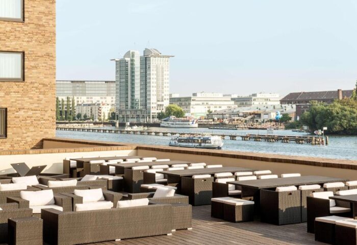Nhow Berlin Hotel gay-friendly terrace view. Tables and chairs overlook the Spree river. A boat cruises by, with Berlin skyline in the background.