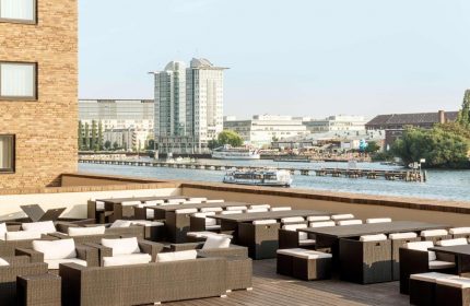 Nhow Berlin Hotel gay-friendly terrace view. Tables and chairs overlook the Spree river. A boat cruises by, with Berlin skyline in the background.