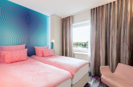 Nhow Berlin Hotel gay-friendly room. Twin beds with pink linens, blue patterned wall, and window view. Modern design for gay travelers.