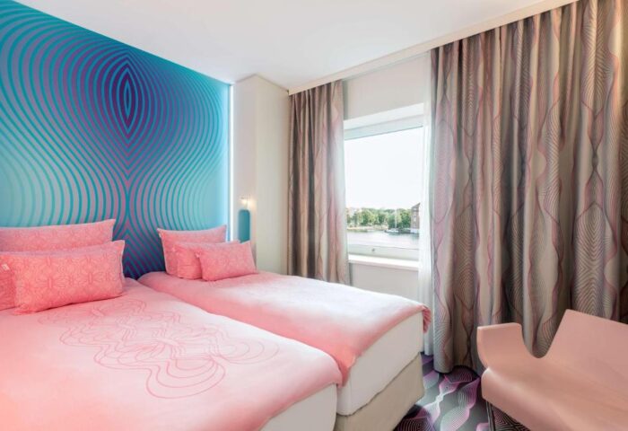 Nhow Berlin Hotel gay-friendly room. Twin beds with pink linens, blue patterned wall, and window view. Modern design for gay travelers.