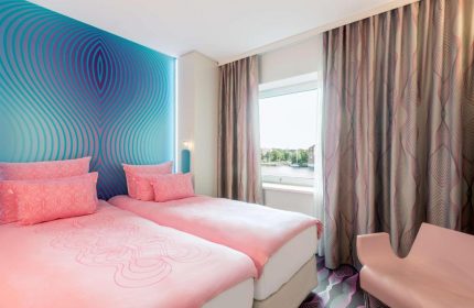 Nhow Berlin Hotel gay-friendly room. Twin beds with pink linens, blue patterned wall, and window view. Modern design for gay travelers.