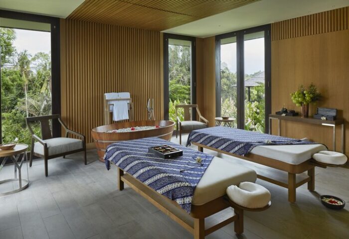 Nirjhara Bali gay-friendly hotel spa. Two massage tables, a wooden bathtub with flowers, and nature views. Luxurious wellness retreat.