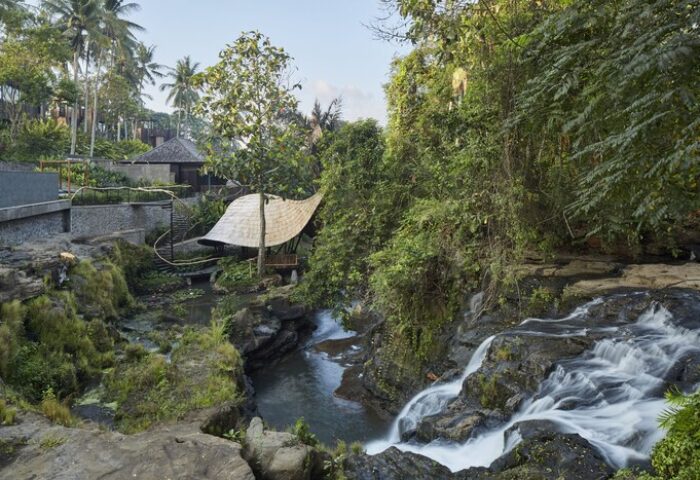 Nirjhara Bali gay-friendly hotel view. Lush greenery surrounds a waterfall cascading into a stream. Thatched roof structure near the water.