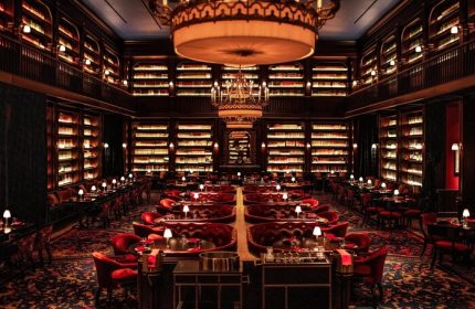 NoMad Las Vegas gay-friendly hotel library restaurant. Elegant dining room with bookshelves, red seating, and chandeliers. Upscale travel.