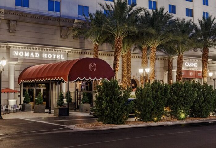 NoMad Las Vegas gay-friendly hotel exterior. Palm trees line the front of the hotel. A red awning covers the entrance. Casino entrance visible.