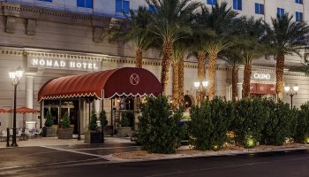 NoMad Las Vegas gay-friendly hotel exterior. Palm trees line the front of the hotel. A red awning covers the entrance. Casino entrance visible.