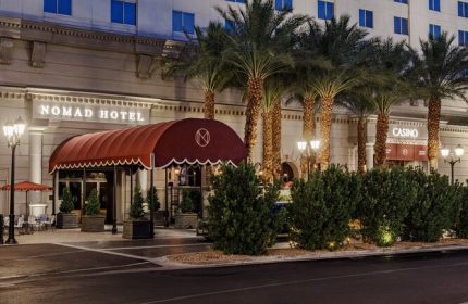 NoMad Las Vegas gay-friendly hotel exterior. Palm trees line the front of the hotel. A red awning covers the entrance. Casino entrance visible.