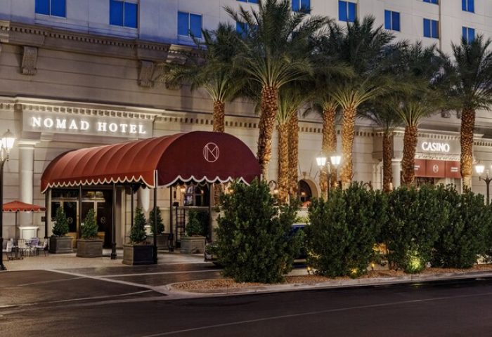 NoMad Las Vegas gay-friendly hotel exterior. Palm trees line the front of the hotel. A red awning covers the entrance. Casino entrance visible.