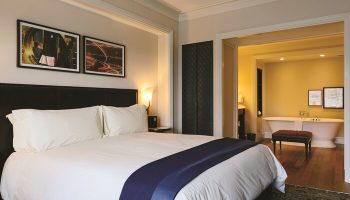 NoMad Las Vegas gay-friendly hotel room: King bed with white linens, dark headboard, and a view into the bathroom with a freestanding tub.