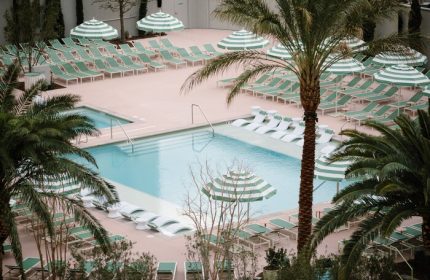 NoMad Las Vegas gay-friendly hotel pool. Palm trees, striped umbrellas, and lounge chairs surround a clear blue pool. Luxury travel.