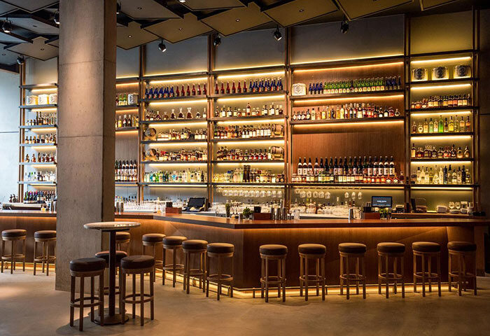 Bar at Nobu Hotel Shoreditch, a gay-friendly, pet-friendly hotel. Shelves display liquor bottles. Stools line the bar. Modern design.