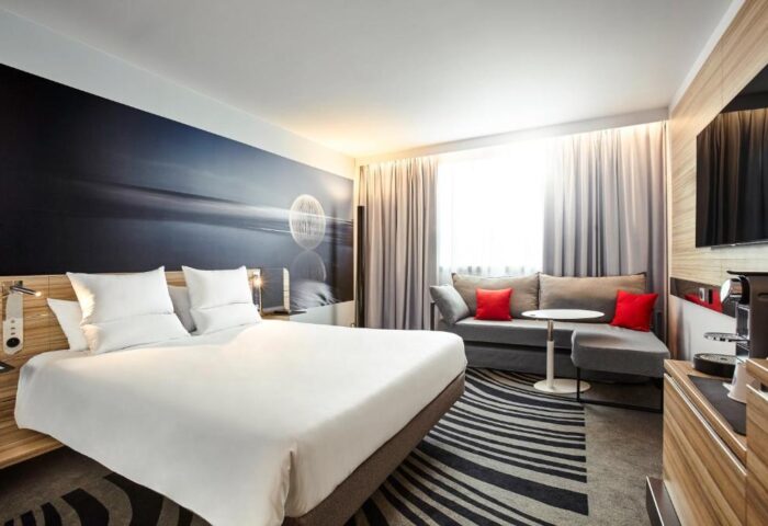 Novotel London Waterloo Hotel gay-friendly room. Modern design with bed, sofa, red pillows, and abstract wall art. Comfortable stay in London.